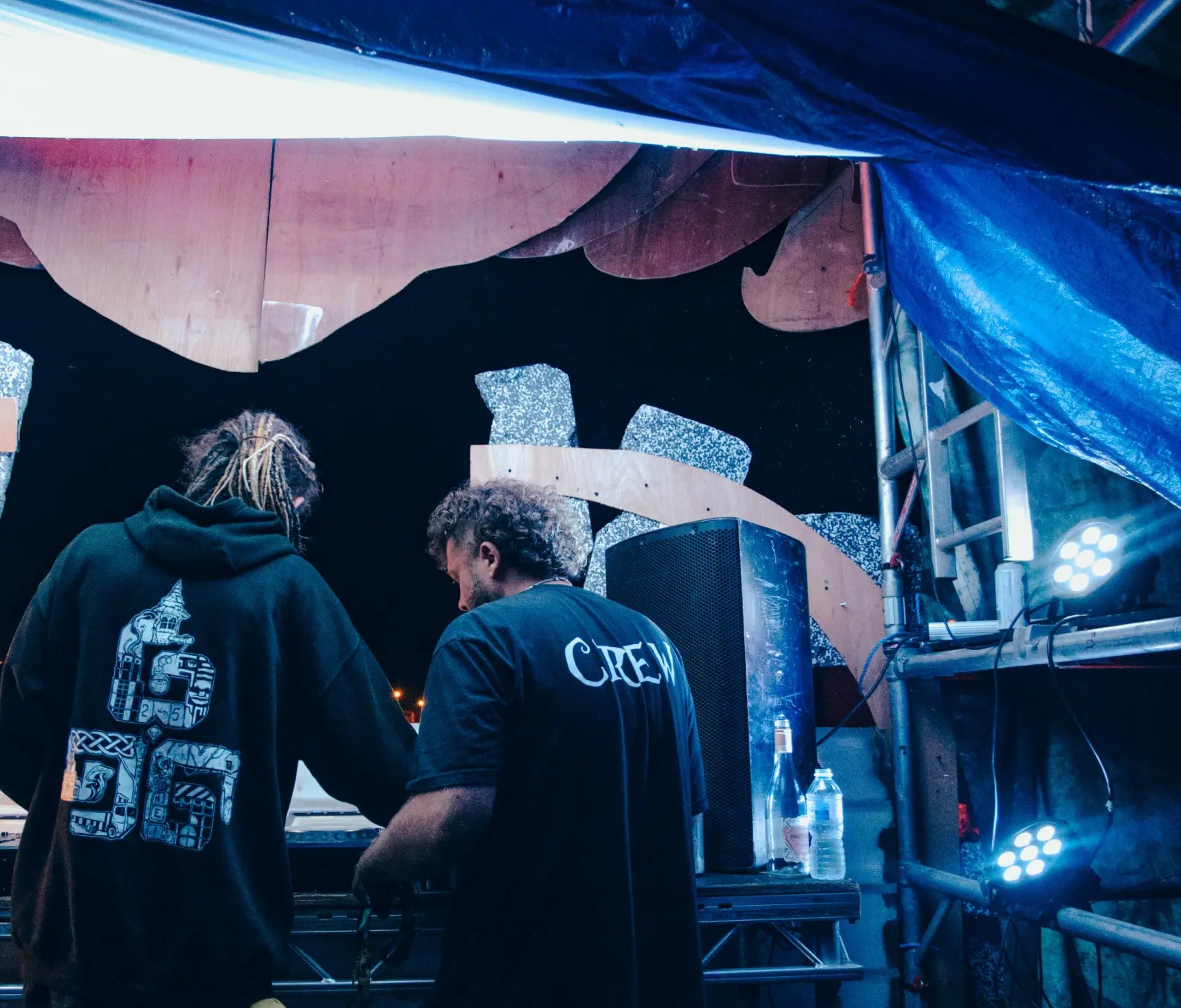 Slipshod crew in printed hoodies watching a set at night, backs to the camera.