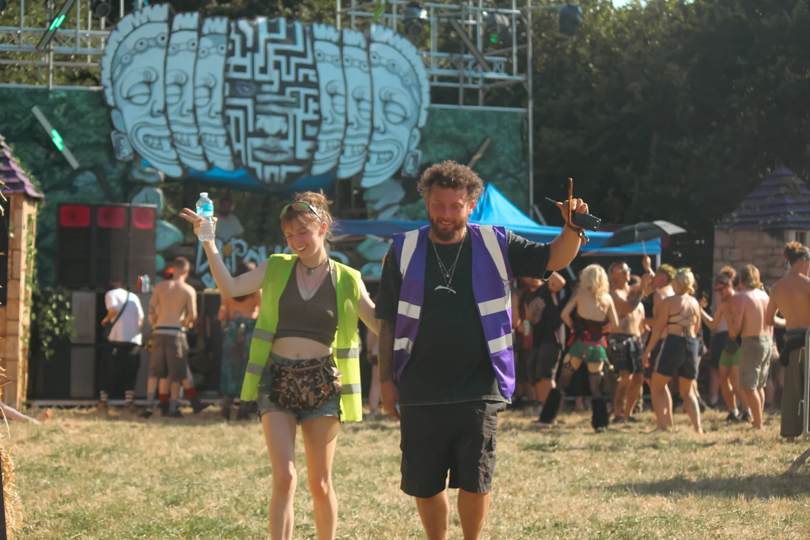 Alex and Abby walking through the festival in hi-vis, radio in hand, The Maw's carved-faces mural and the crowd behind them